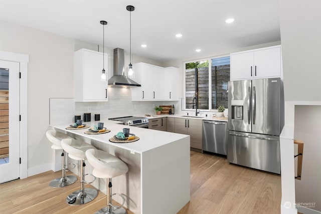 kitchen with kitchen peninsula, decorative light fixtures, wall chimney range hood, appliances with stainless steel finishes, and light wood-type flooring