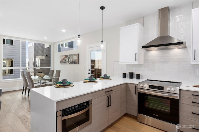 kitchen with wall chimney exhaust hood, light wood-type flooring, decorative backsplash, hanging light fixtures, and appliances with stainless steel finishes