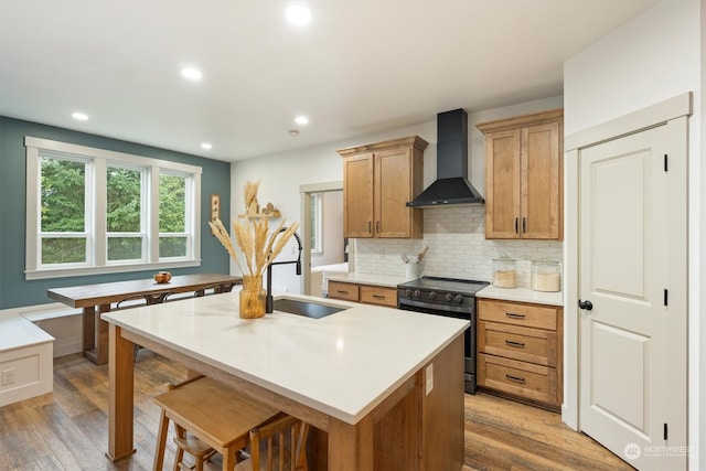 kitchen featuring wall chimney exhaust hood, hardwood / wood-style floors, a kitchen island with sink, and black range with electric stovetop