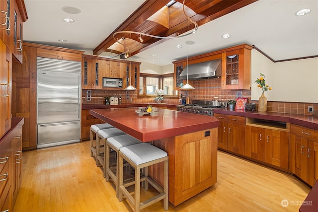 kitchen with wall chimney exhaust hood, light hardwood / wood-style floors, a kitchen breakfast bar, decorative backsplash, and built in appliances