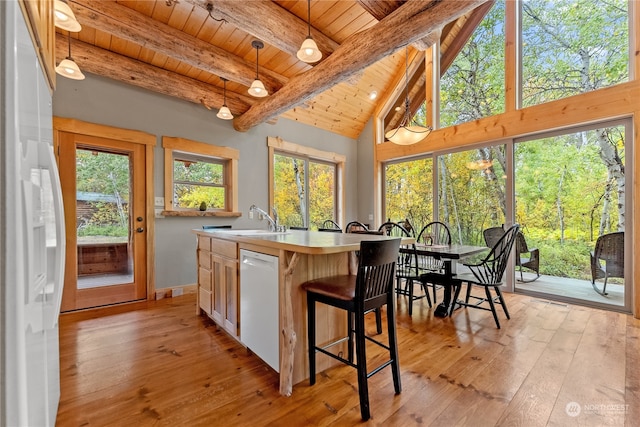 kitchen with pendant lighting, beamed ceiling, white appliances, a kitchen island with sink, and a wealth of natural light