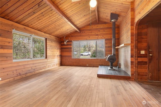 unfurnished living room featuring wooden walls, wood ceiling, a wood stove, light hardwood / wood-style floors, and vaulted ceiling with beams