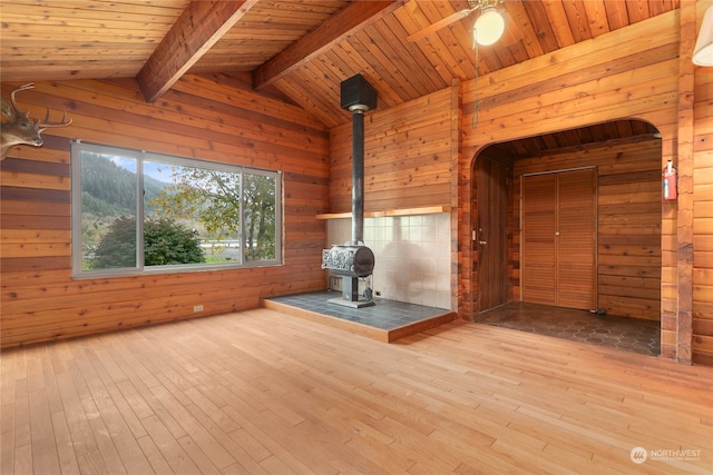 unfurnished living room featuring wood walls, a wood stove, lofted ceiling with beams, wooden ceiling, and light wood-type flooring