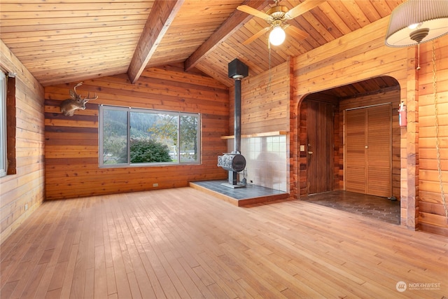 unfurnished living room featuring wood walls, light hardwood / wood-style floors, wood ceiling, a wood stove, and vaulted ceiling with beams