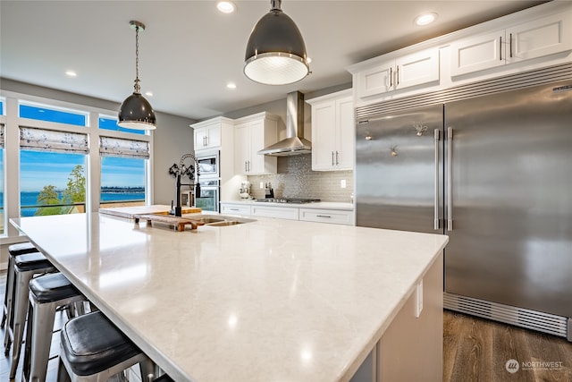 kitchen with built in appliances, white cabinetry, wall chimney range hood, and hanging light fixtures