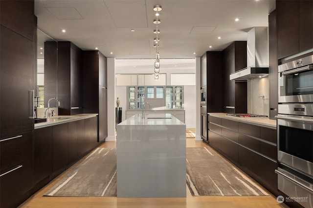 kitchen featuring an island with sink, pendant lighting, wall chimney exhaust hood, and light hardwood / wood-style floors