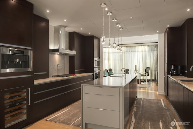 kitchen featuring wall chimney exhaust hood, a kitchen island with sink, decorative light fixtures, wine cooler, and sink