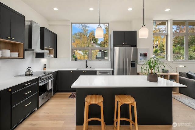 kitchen with wall chimney exhaust hood, sink, pendant lighting, appliances with stainless steel finishes, and a kitchen bar