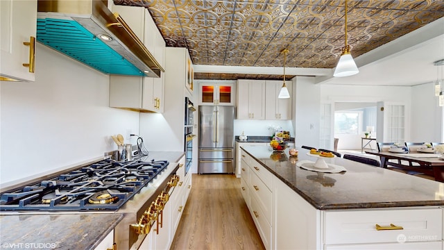 kitchen featuring wall chimney exhaust hood, dark stone countertops, decorative light fixtures, white cabinets, and appliances with stainless steel finishes