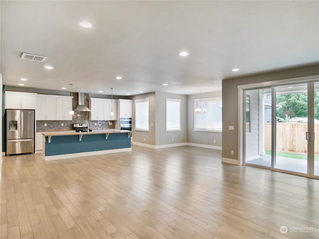 kitchen featuring white cabinetry, stainless steel appliances, wall chimney exhaust hood, decorative light fixtures, and a center island with sink