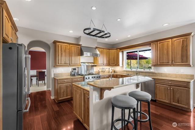 kitchen with wall chimney range hood, appliances with stainless steel finishes, a center island, a breakfast bar, and dark hardwood / wood-style floors