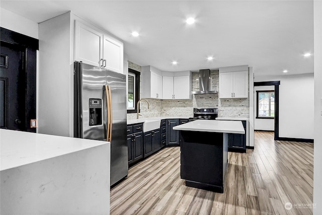 kitchen with wall chimney range hood, a kitchen island, appliances with stainless steel finishes, white cabinetry, and light hardwood / wood-style floors