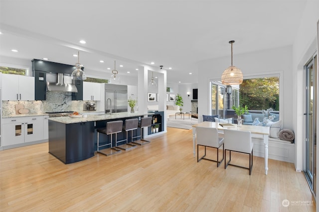 kitchen with white cabinetry, backsplash, a large island with sink, light hardwood / wood-style floors, and decorative light fixtures