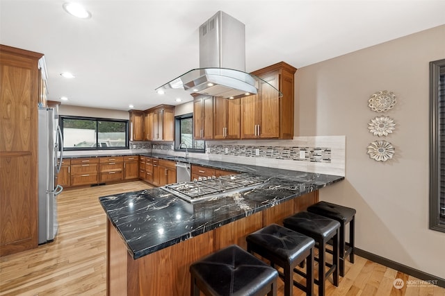 kitchen featuring kitchen peninsula, appliances with stainless steel finishes, island range hood, light hardwood / wood-style flooring, and a breakfast bar area