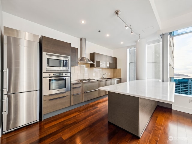 kitchen featuring decorative backsplash, wall chimney exhaust hood, dark wood-type flooring, light stone countertops, and appliances with stainless steel finishes