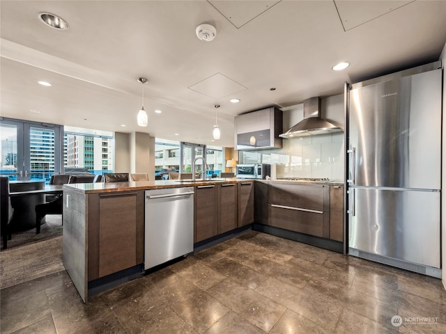 kitchen with sink, kitchen peninsula, stainless steel appliances, wall chimney exhaust hood, and decorative light fixtures