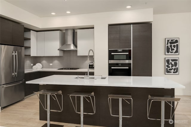 kitchen featuring wall chimney range hood, stainless steel appliances, a center island with sink, and a kitchen bar