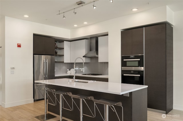 kitchen with black appliances, light hardwood / wood-style floors, wall chimney exhaust hood, a breakfast bar, and a kitchen island with sink