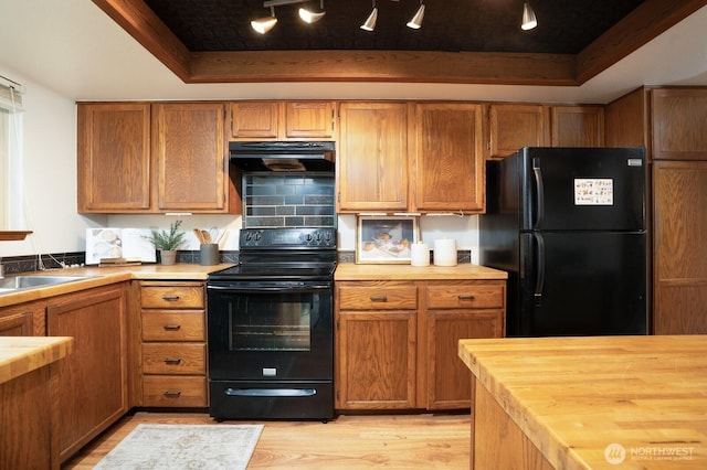 kitchen with wooden counters, a tray ceiling, black appliances, light wood-style floors, and under cabinet range hood