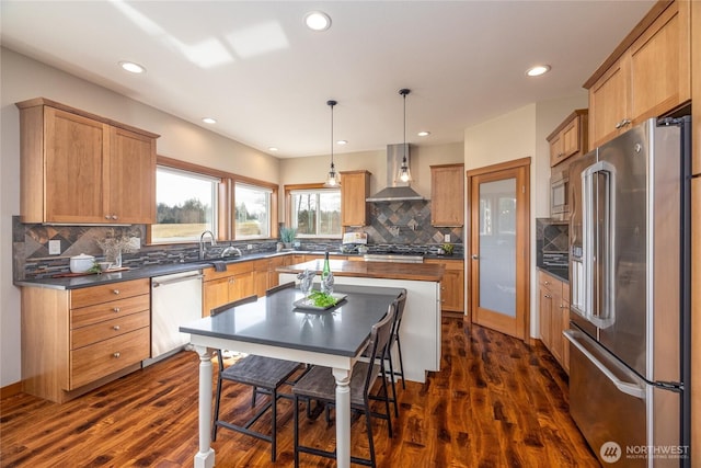 kitchen with dark wood-type flooring, appliances with stainless steel finishes, wall chimney range hood, dark countertops, and decorative light fixtures