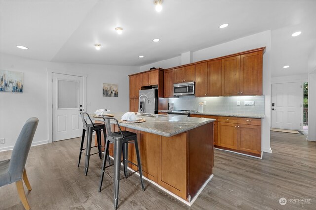 kitchen with tasteful backsplash, appliances with stainless steel finishes, light stone counters, hardwood / wood-style flooring, and a center island with sink