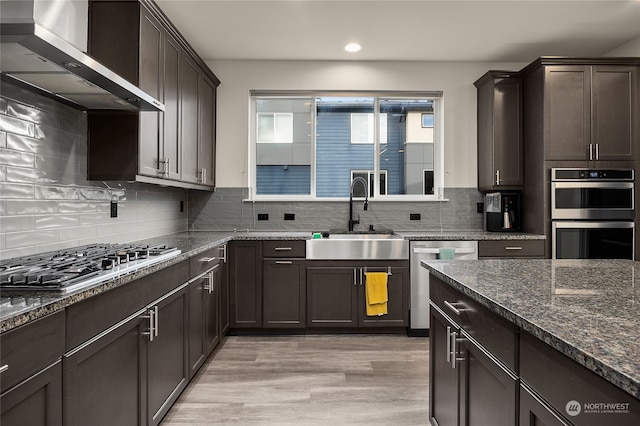 kitchen with appliances with stainless steel finishes, tasteful backsplash, wall chimney exhaust hood, sink, and light hardwood / wood-style flooring