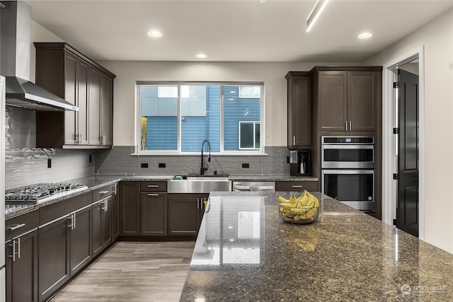 kitchen with appliances with stainless steel finishes, dark stone counters, sink, wall chimney range hood, and light hardwood / wood-style floors