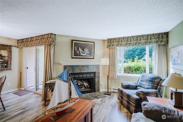 living room featuring a tiled fireplace, a textured ceiling, baseboard heating, and light hardwood / wood-style flooring