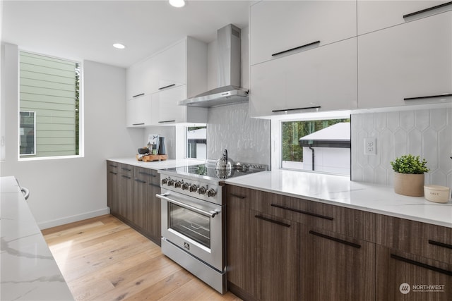 kitchen featuring high end range, white cabinetry, light hardwood / wood-style floors, wall chimney exhaust hood, and light stone counters