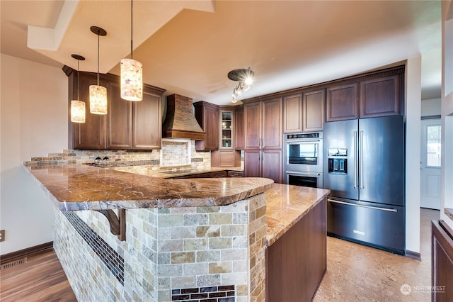 kitchen featuring backsplash, kitchen peninsula, stainless steel appliances, decorative light fixtures, and premium range hood