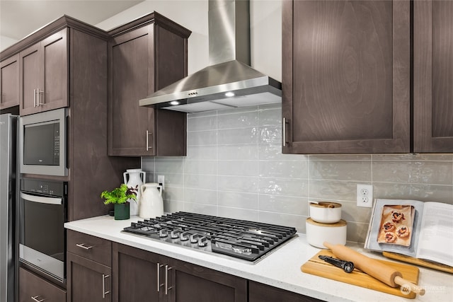 kitchen with dark brown cabinetry, stainless steel appliances, decorative backsplash, and wall chimney range hood