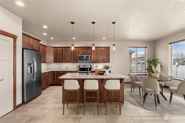 kitchen featuring a kitchen bar, appliances with stainless steel finishes, light stone counters, a kitchen island with sink, and hanging light fixtures