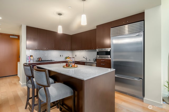 kitchen with dark brown cabinetry, pendant lighting, built in appliances, decorative backsplash, and light hardwood / wood-style flooring