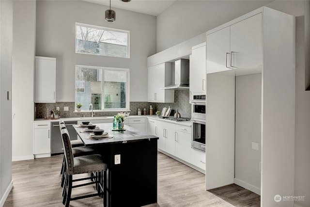 kitchen featuring a sink, wall chimney range hood, appliances with stainless steel finishes, light wood-type flooring, and decorative backsplash