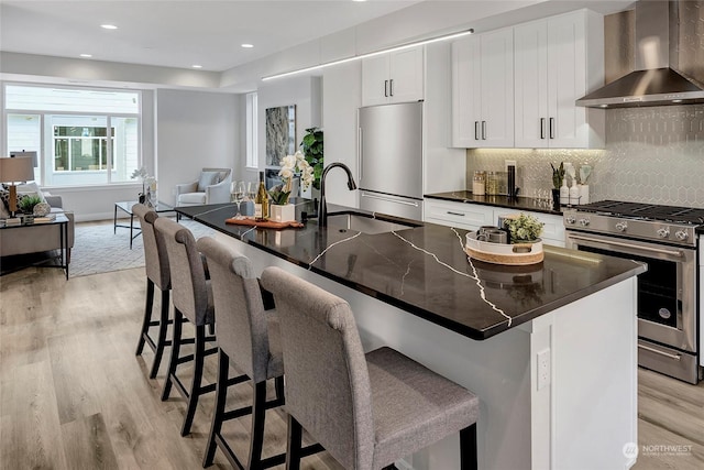 kitchen with a breakfast bar area, stainless steel appliances, an island with sink, and wall chimney exhaust hood