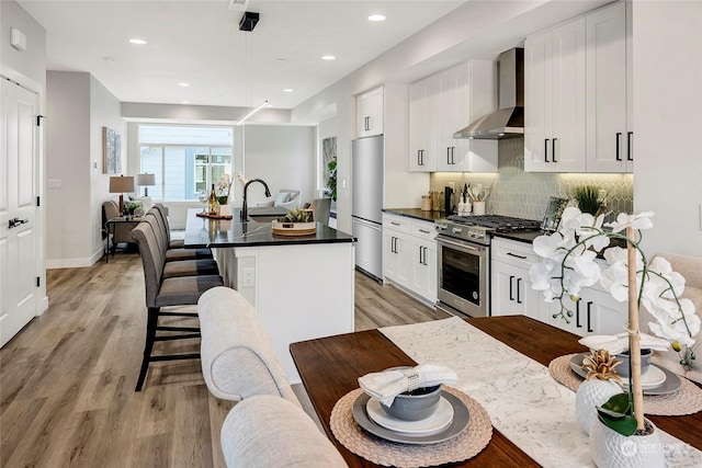 kitchen with wall chimney range hood, a breakfast bar area, white cabinetry, stainless steel appliances, and an island with sink