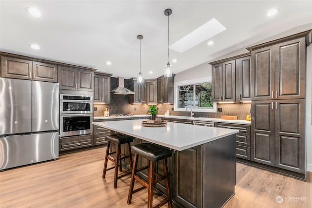 kitchen with light hardwood / wood-style floors, wall chimney range hood, vaulted ceiling with skylight, a kitchen island, and appliances with stainless steel finishes