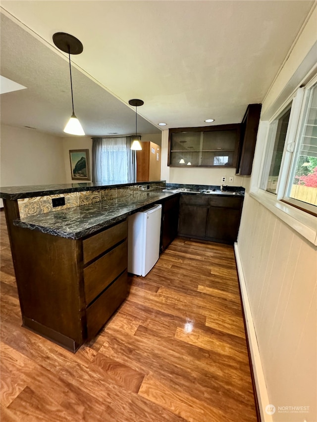 kitchen featuring kitchen peninsula, dark brown cabinets, dark stone counters, dark hardwood / wood-style floors, and decorative light fixtures