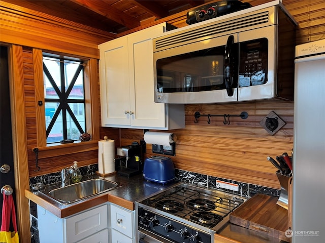 kitchen featuring stainless steel appliances, wooden walls, sink, beamed ceiling, and white cabinets