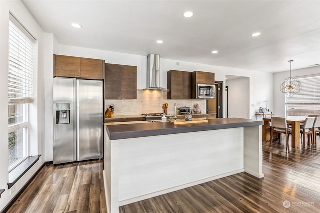 kitchen with wall chimney exhaust hood, dark wood-type flooring, an island with sink, pendant lighting, and appliances with stainless steel finishes