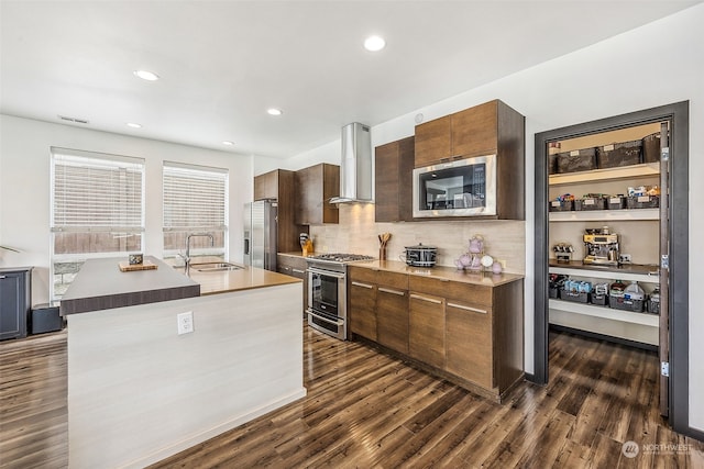 kitchen with wall chimney range hood, sink, stainless steel appliances, dark hardwood / wood-style floors, and a kitchen island with sink