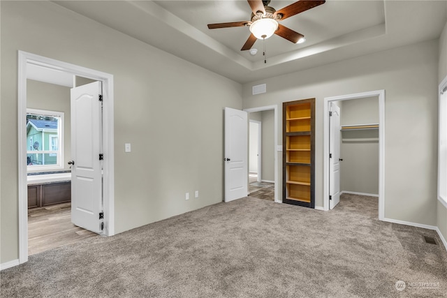 unfurnished bedroom with a spacious closet, a tray ceiling, a closet, ceiling fan, and light colored carpet