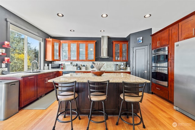 kitchen with appliances with stainless steel finishes, light wood-style floors, a sink, a kitchen island, and wall chimney exhaust hood