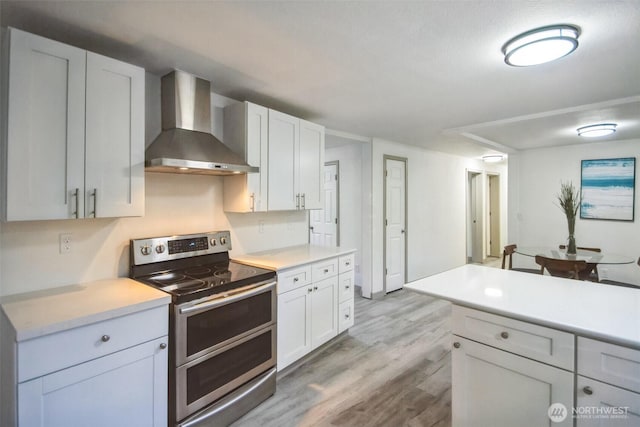 kitchen featuring light wood finished floors, wall chimney range hood, double oven range, light countertops, and white cabinetry