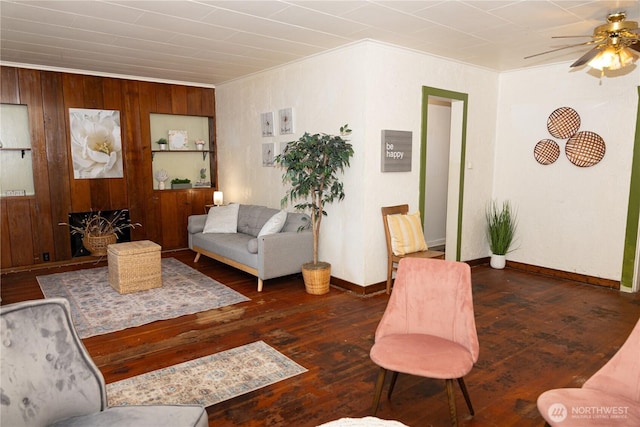 living area featuring crown molding, a ceiling fan, baseboards, wooden walls, and dark wood-style flooring