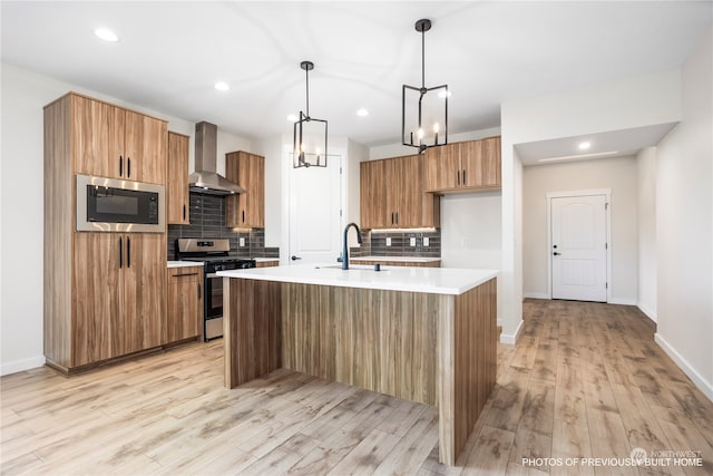 kitchen with stainless steel appliances, light hardwood / wood-style floors, an island with sink, wall chimney range hood, and pendant lighting