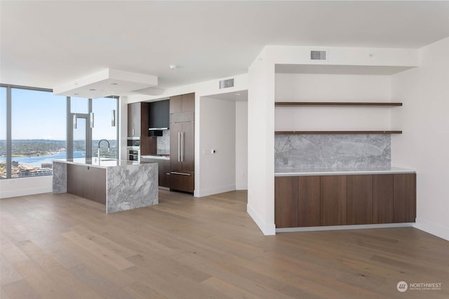 kitchen featuring light hardwood / wood-style flooring, a water view, a center island with sink, decorative backsplash, and dark brown cabinets