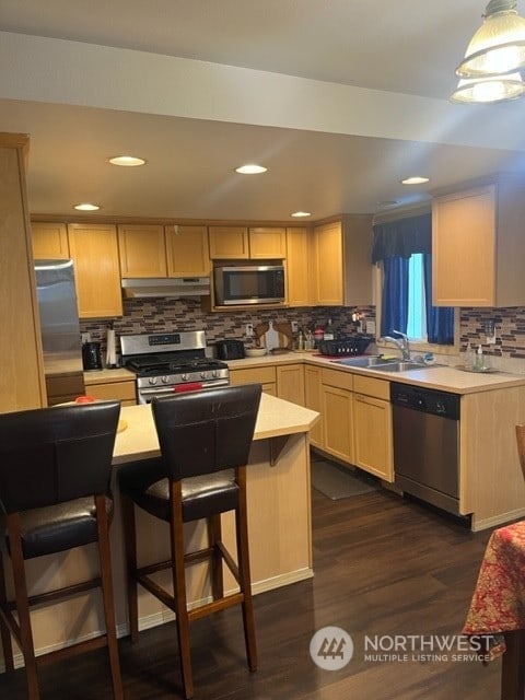 kitchen with stainless steel appliances, light brown cabinets, a breakfast bar area, backsplash, and dark hardwood / wood-style flooring