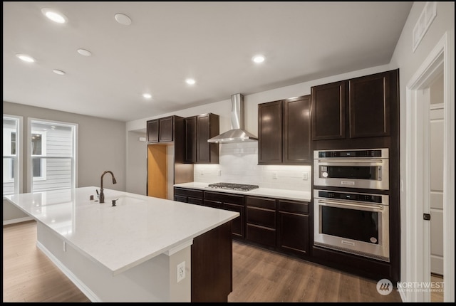 kitchen with hardwood / wood-style floors, wall chimney exhaust hood, a kitchen island with sink, and appliances with stainless steel finishes