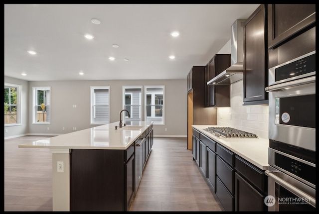 kitchen featuring a center island with sink, stainless steel appliances, wall chimney range hood, sink, and light hardwood / wood-style flooring